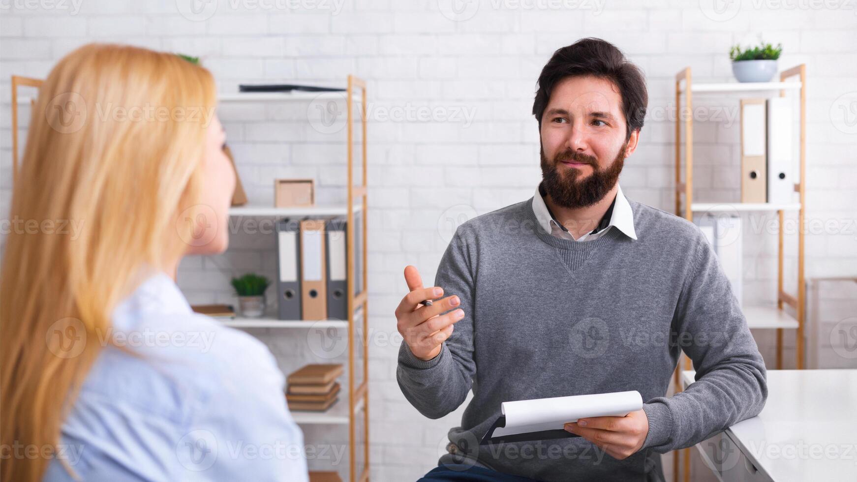 A man with a beard is seated across from a woman, engaged in conversation. He holds a document, possibly a resume, gesturing as he speaks, suggesting a job interview scenario or therapeutic session photo
