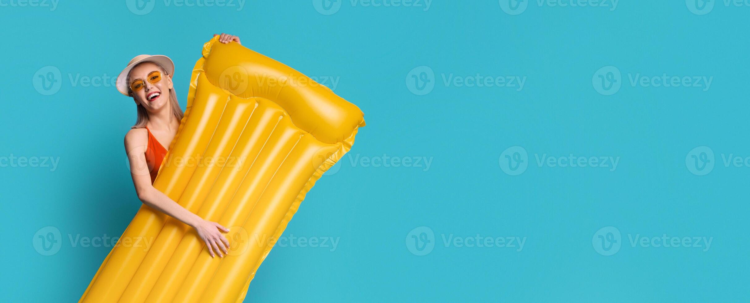 A young woman in a red swimsuit and a white sunhat smiles and holds a yellow inflatable raft in front of a solid blue background. The woman is looking at the camera, copy space photo