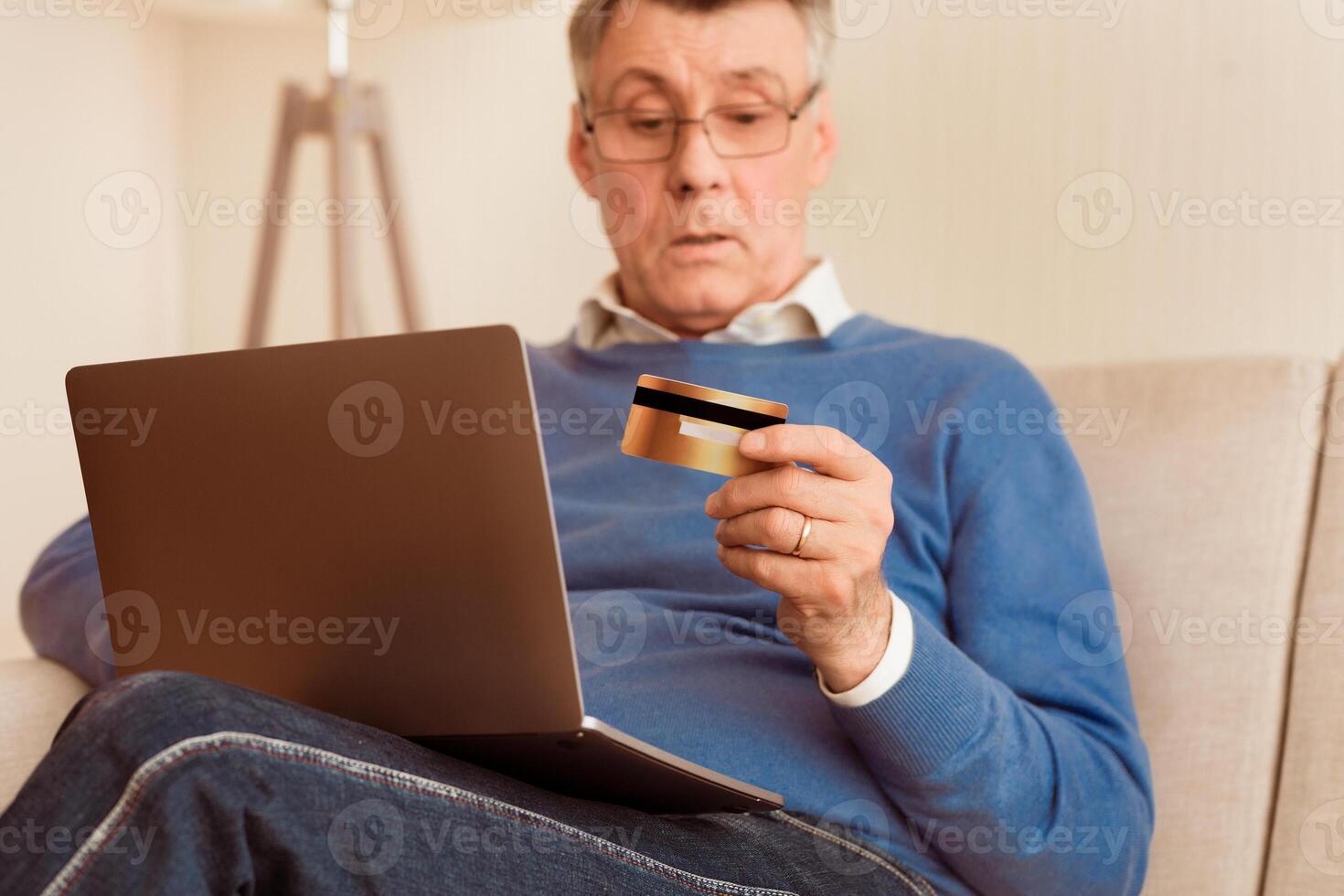 Senior Man Using Credit Card And Laptop Shopping Online Sitting On Couch At Home. Selective Focus photo
