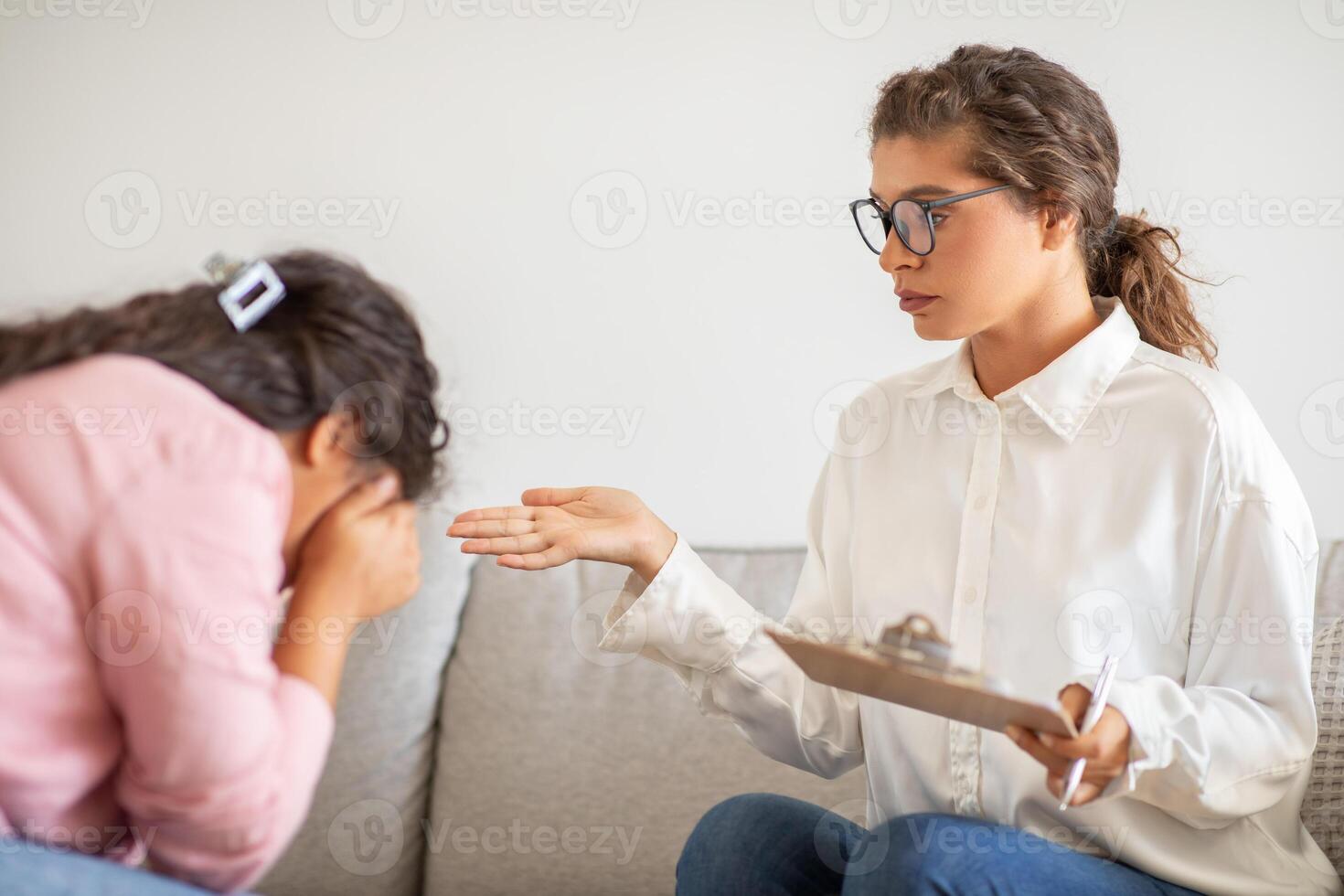 A therapist, wearing a white shirt and glasses, sits on a couch and gestures with her right hand while speaking to a patient. The patient, with their head down, appears to be crying or distressed photo