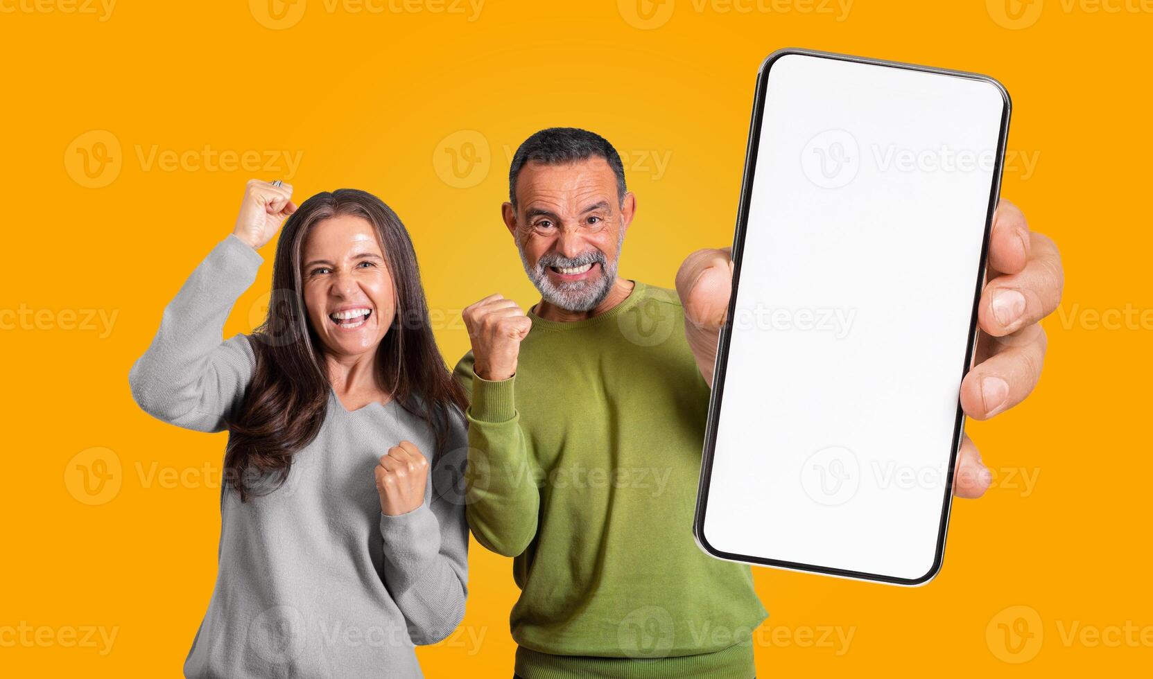 Mature man and woman are standing in front of yellow background, both looking at the camera and smiling excitedly with their fists raised in the air, holding a smartphone with a blank white screen photo
