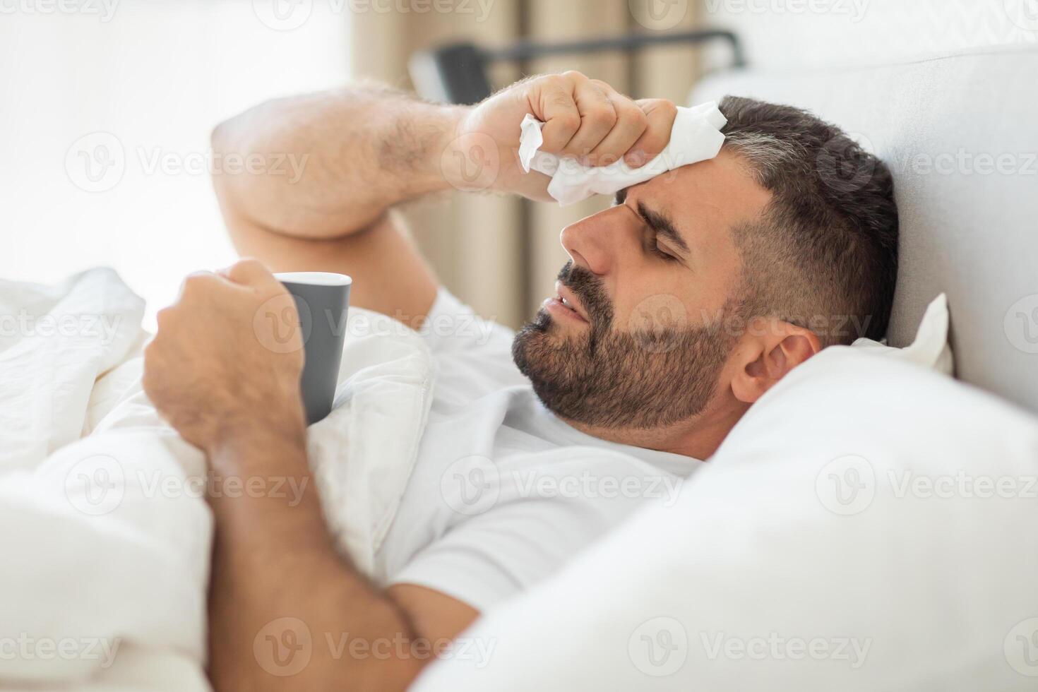 A man lies in bed with a tissue in his hand, wiping his forehead. He appears to be sick or unwell, as he holds a mug and has a tired expression on his face. He is covered by white bedsheets photo