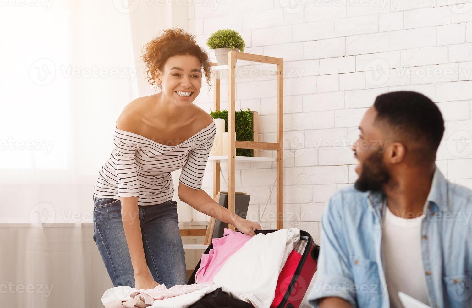 African American couple in a room, unpacking clothes from suitcases. The man is folding shirts while the woman hangs dresses in the closet, back from trip photo