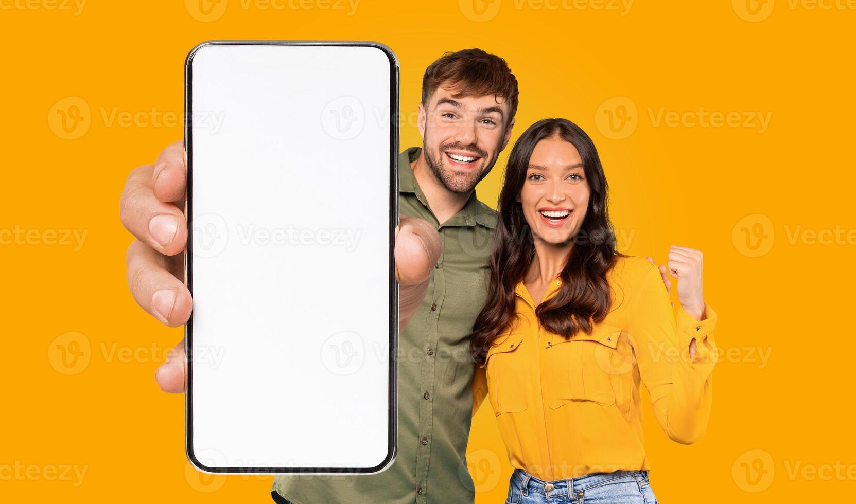 A happy couple is holding a smartphone with a blank white screen, and they are both pointing at the screen with smiles on their faces. They are standing in front of a bright yellow background. photo