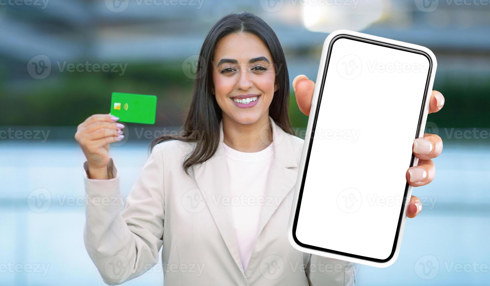 A woman is standing and holding up a smart phone in one hand and a credit card in the other hand. She appears to be engaged in a transaction or making a purchase. photo