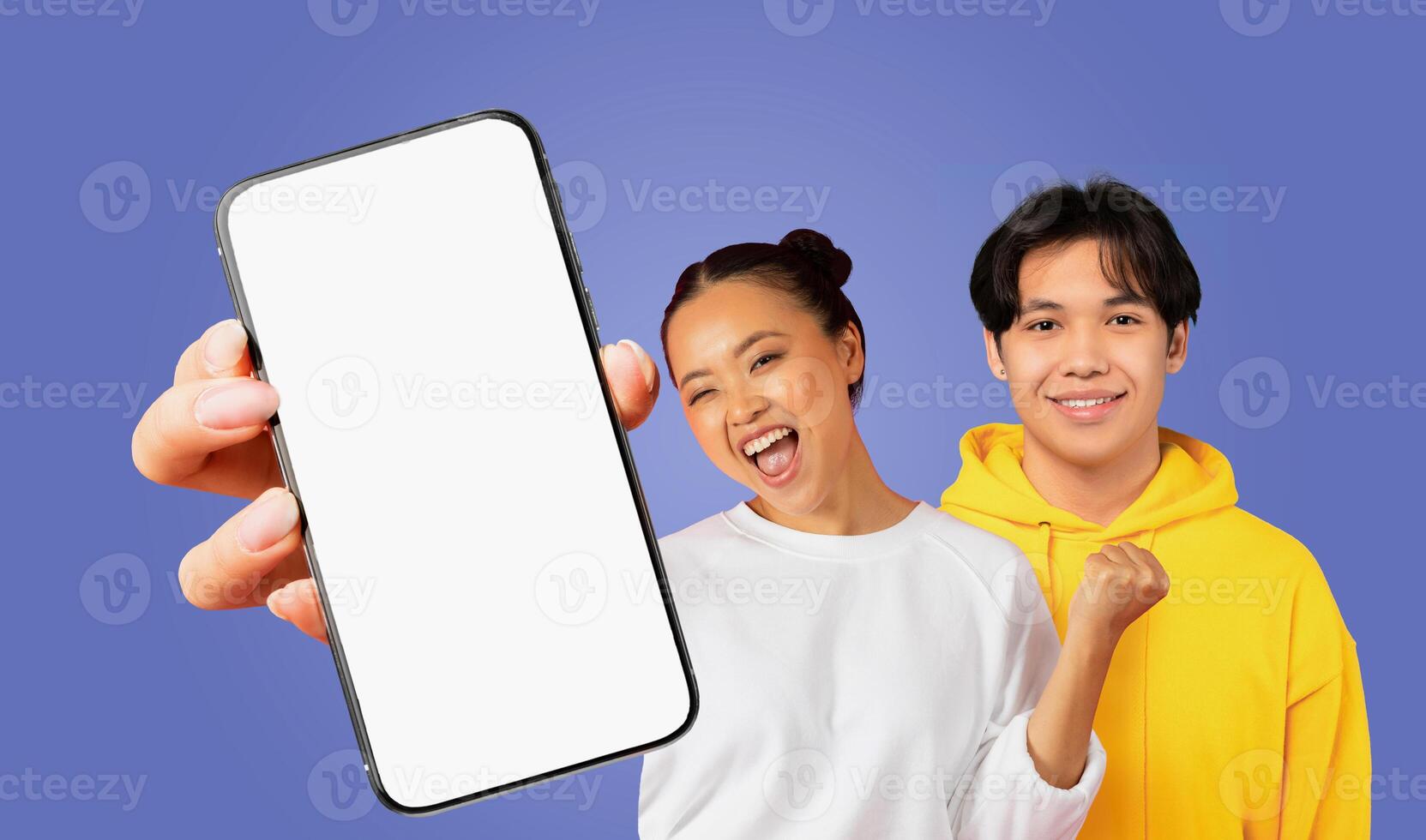 Asian couple standing side-by-side in front of a plain purple background. The woman is smiling widely and looking at the camera while holding a smartphone with a blank white screen photo