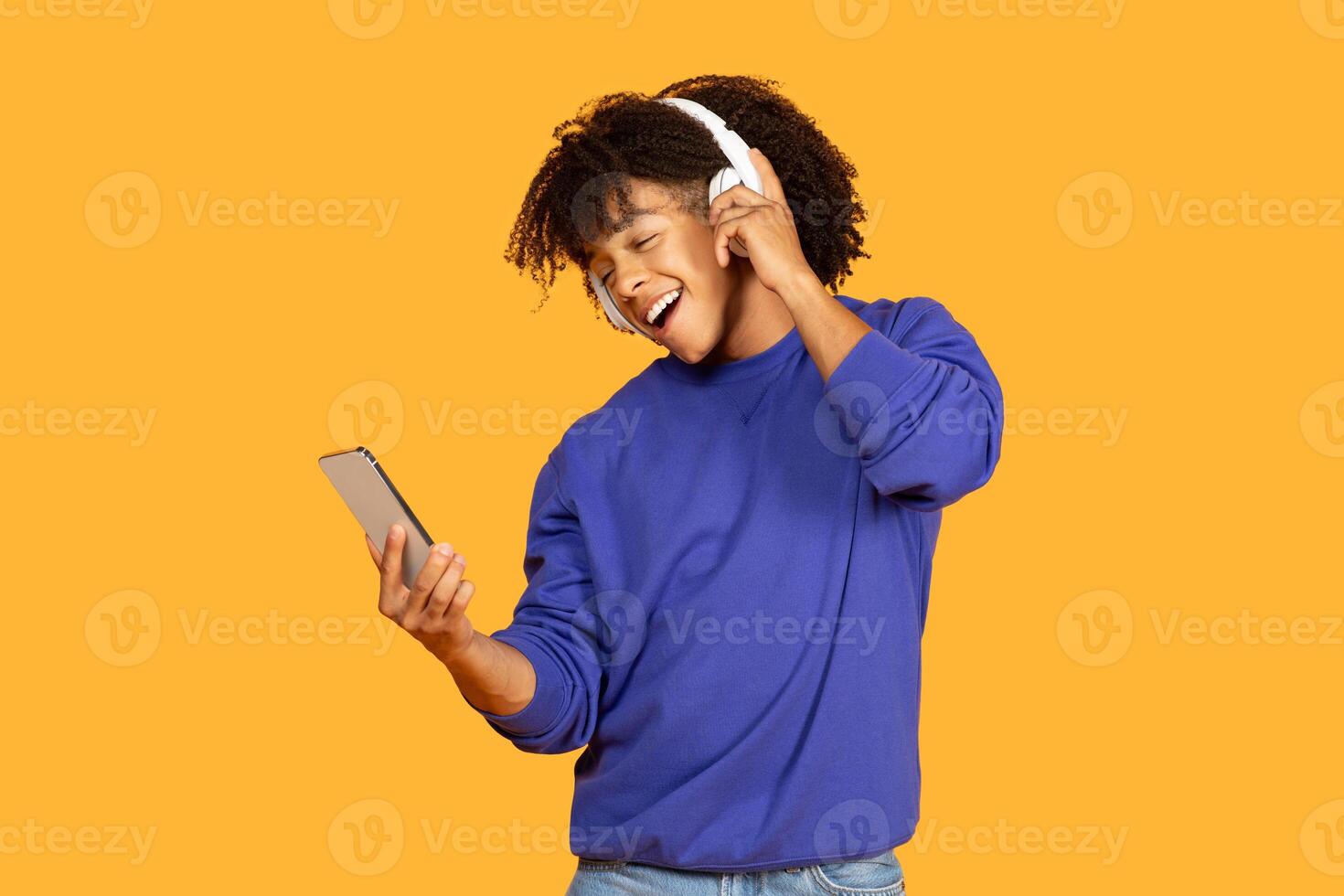 A young man with curly hair joyfully listens to music through headphones while holding a smartphone. The bright yellow background enhances the vibrant and lively atmosphere photo