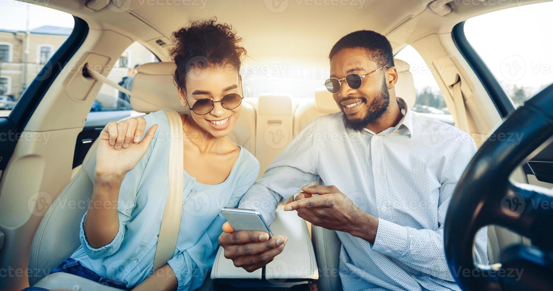 A young black couple, wearing sunglasses and casual clothes, are sitting in the back seat of a car and looking at a smartphone. They are smiling and appear to be enjoying each others company photo