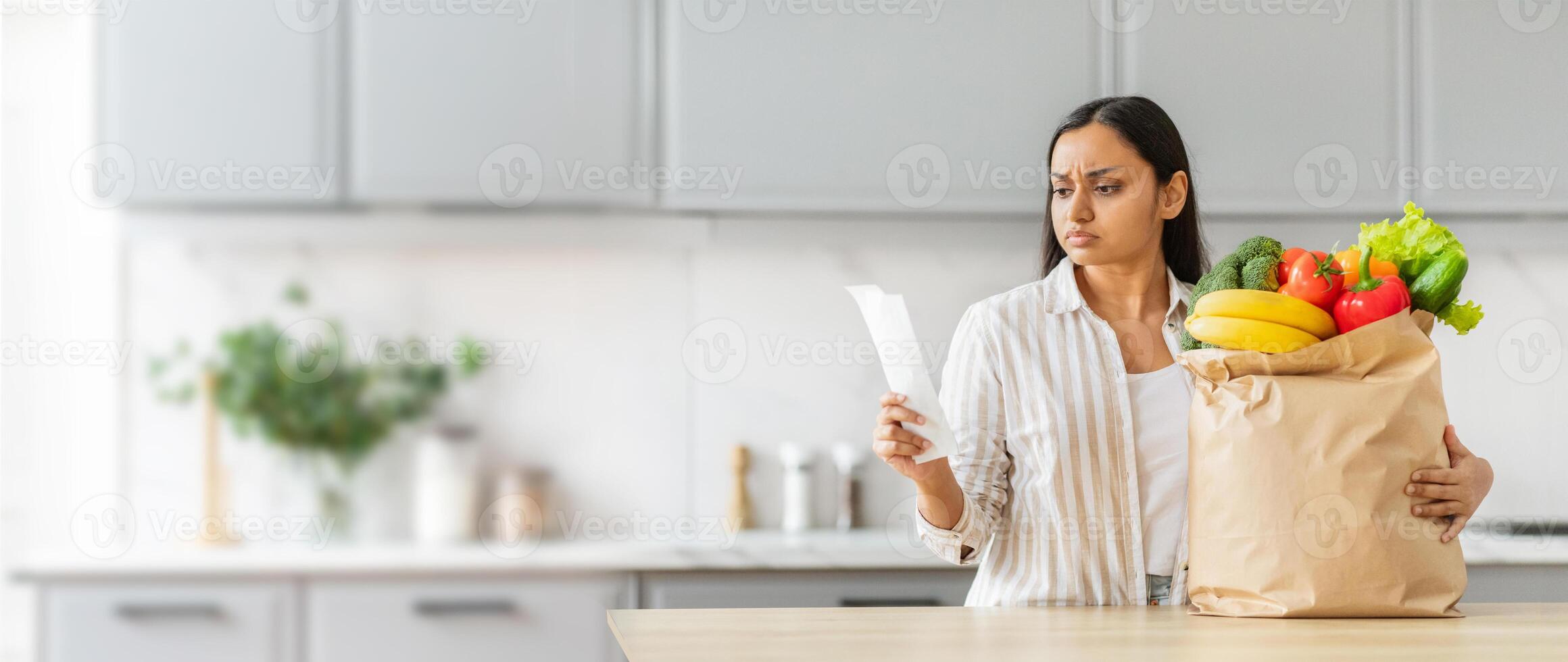Indian woman stands in her kitchen, looking at a receipt in her hand with a shocked expression. She holding a paper bag filled with fresh goods, copy space photo