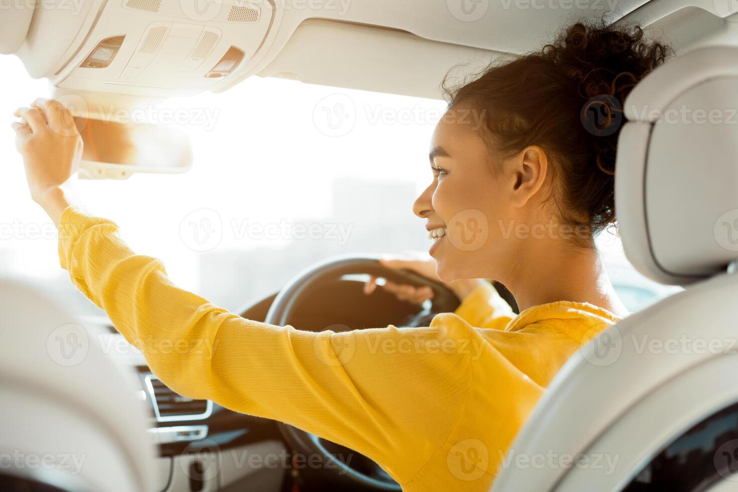 This image shows a young woman with curly hair sitting in the drivers seat of a car. She is wearing a yellow blouse and smiling as she adjusts the rearview mirror above her head. photo