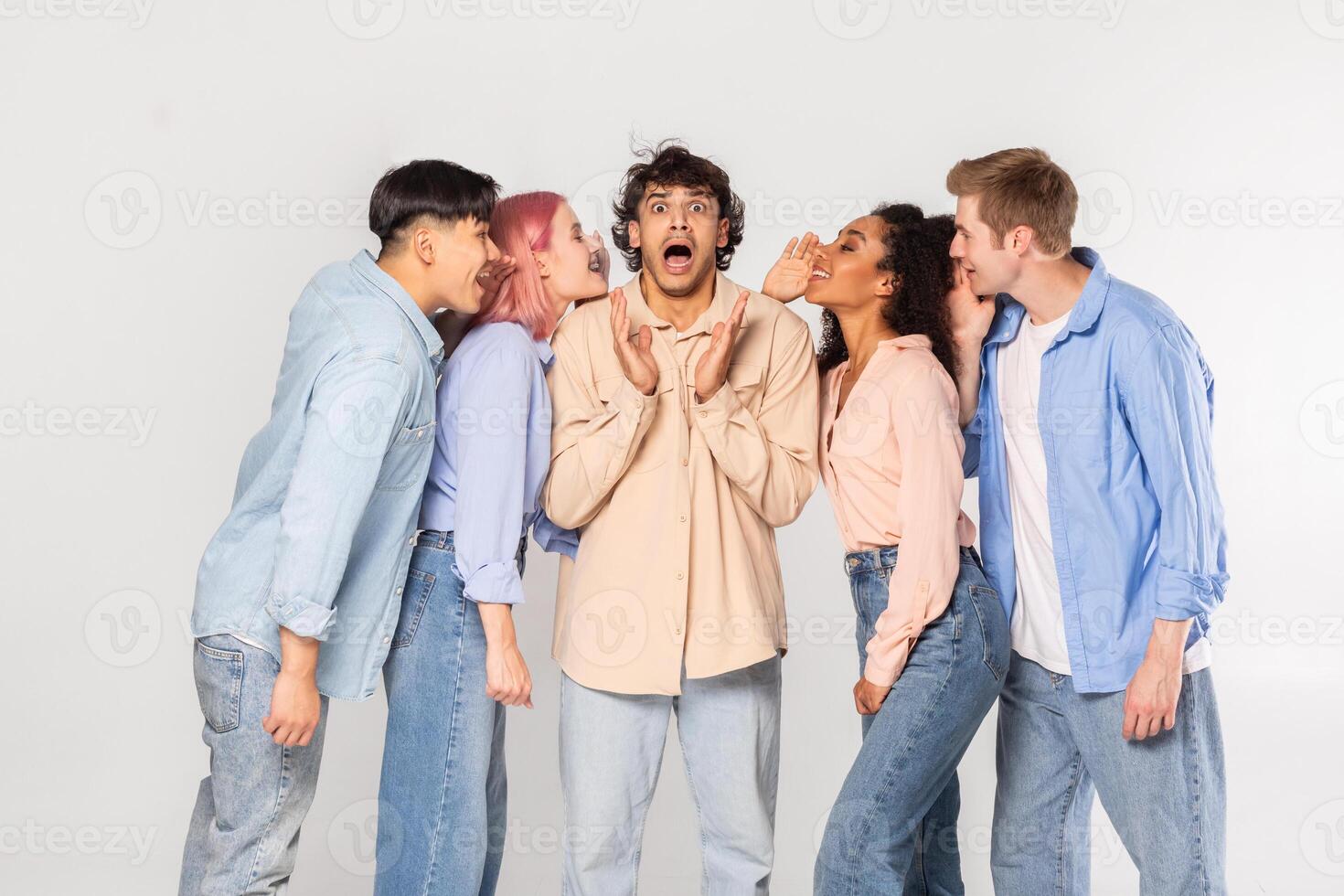 A group of four friends are gathered around a man in a light brown shirt. The friends are all whispering secrets to the man, who looks surprised and confused. photo