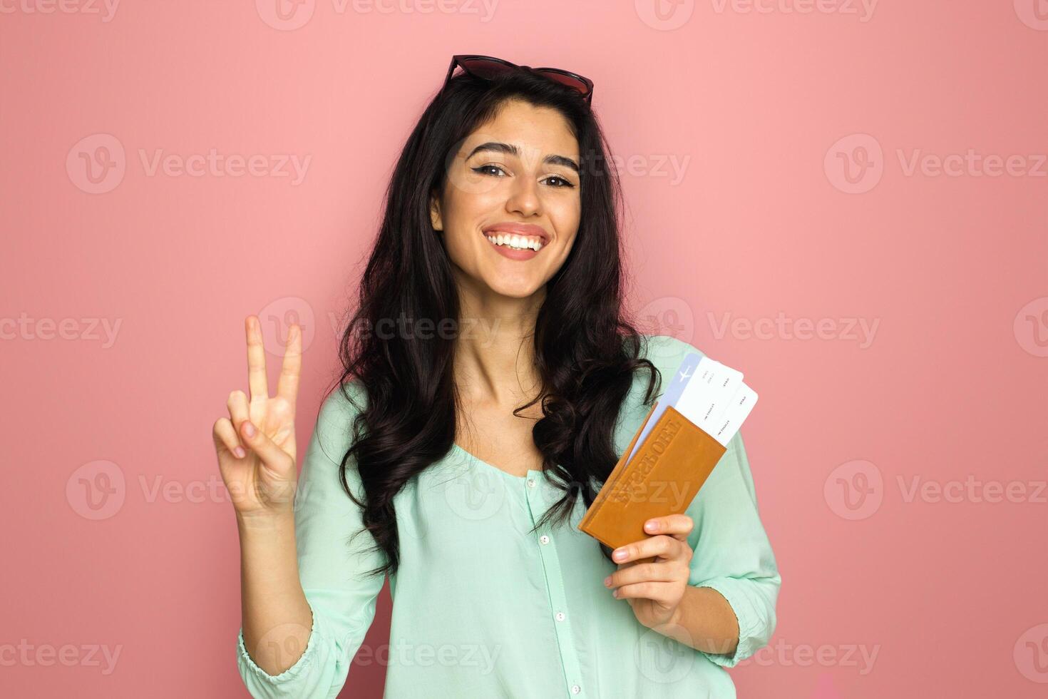 A cheerful young middle eastern woman, ready for travel, stands against a pink background holding her passport and boarding pass, flashing a peace sign with a beaming smile. photo