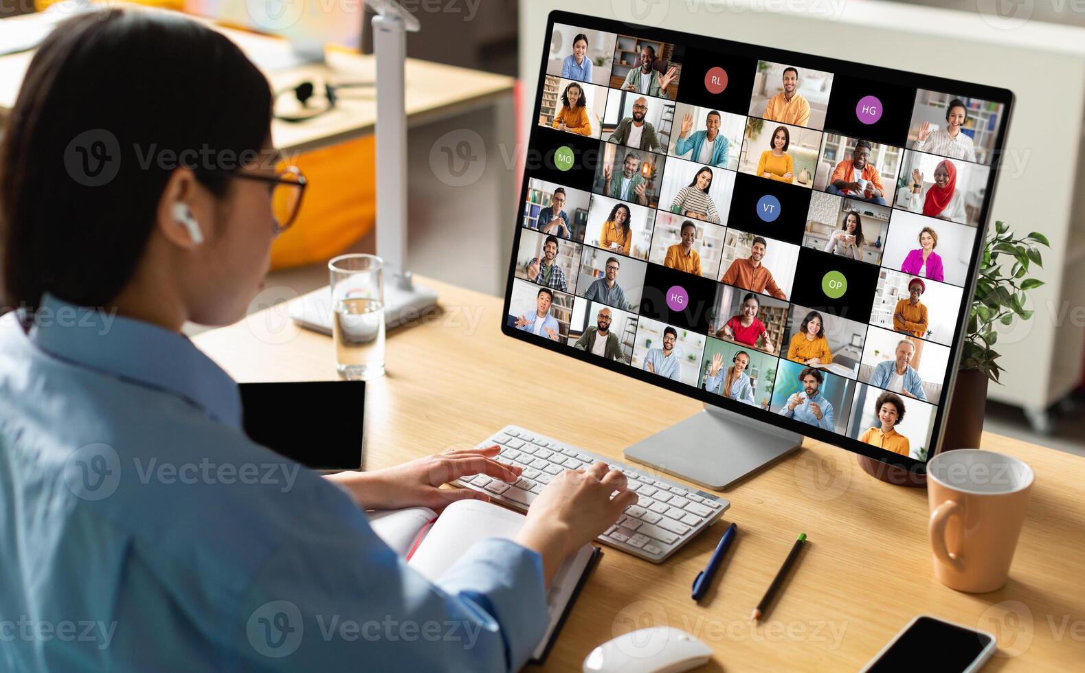 A woman wearing earbuds sits at her desk in front of a computer monitor displaying a conference call with many participants. She is actively participating in the meeting photo