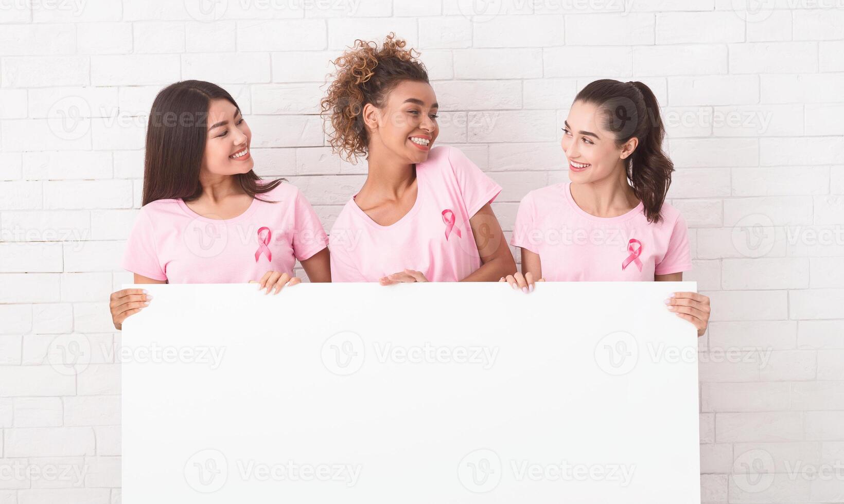 Three Multiethnic Girls In Pink Breast Cancer T-Shirts Holding Blank White Board Against Brick Wall. Mockup photo