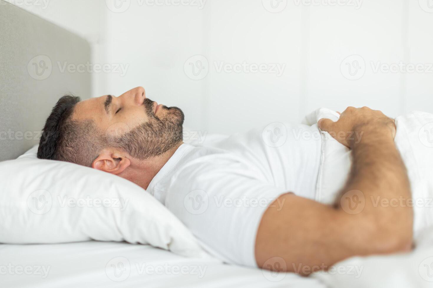 A man with a beard is sleeping on his side in a bed. He is lying on a white pillow, with a white sheet pulled up to his chest. The room is bright and the bed is white. photo