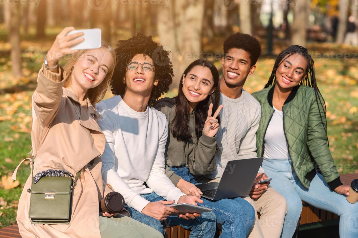 Cheerful international teenagers sitting on bench and taking group selfie on smartphone photo