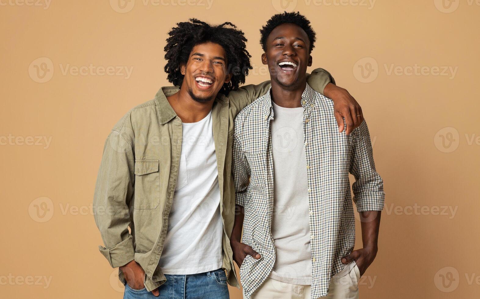 Portrait Of Two Happy Black Guys Embracing While Posing Over Beige Background, Cheerful Young African American Friends In Stylish Clothes Having Fun In Studio, Laughing At Camera, Copy Space photo
