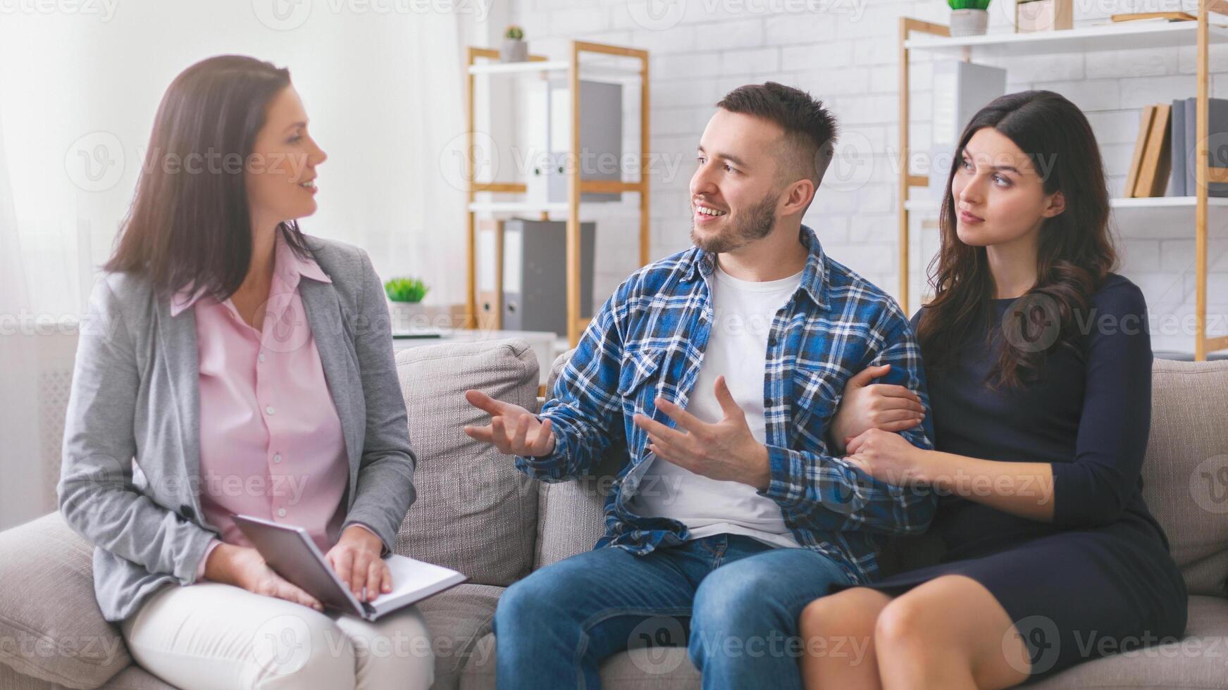 A young couple sits on a couch in a modern office setting, while a therapist listens attentively. The man is speaking with his hands, while the woman sits quietly beside him. photo