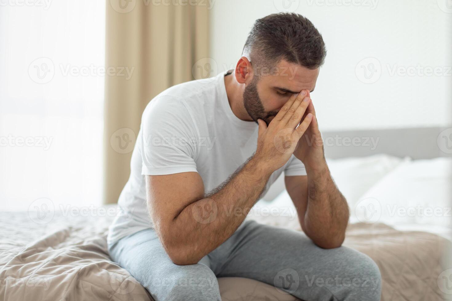 A man wearing a white t-shirt and gray pants is sitting on the edge of a bed. He has his hands on his face and is looking down, appearing to be in deep thought. photo