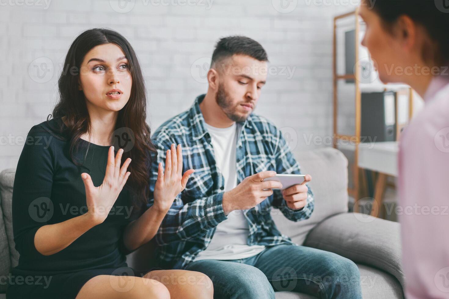A young couple is sitting on a couch in a therapists office. The woman is talking to the therapist while the man is looking at his phone. The woman is visibly upset. photo
