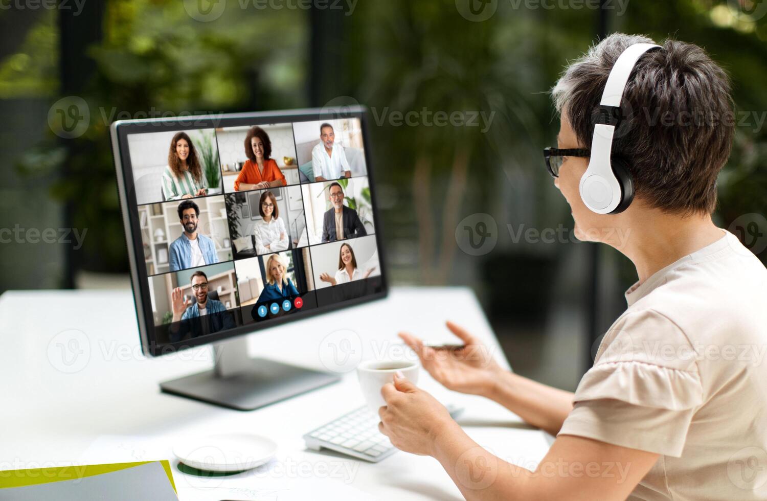 Woman is sitting at a desk, focusing on a computer screen. She is wearing headphones, possibly listening to music or participating in a call. The room is well-lit photo