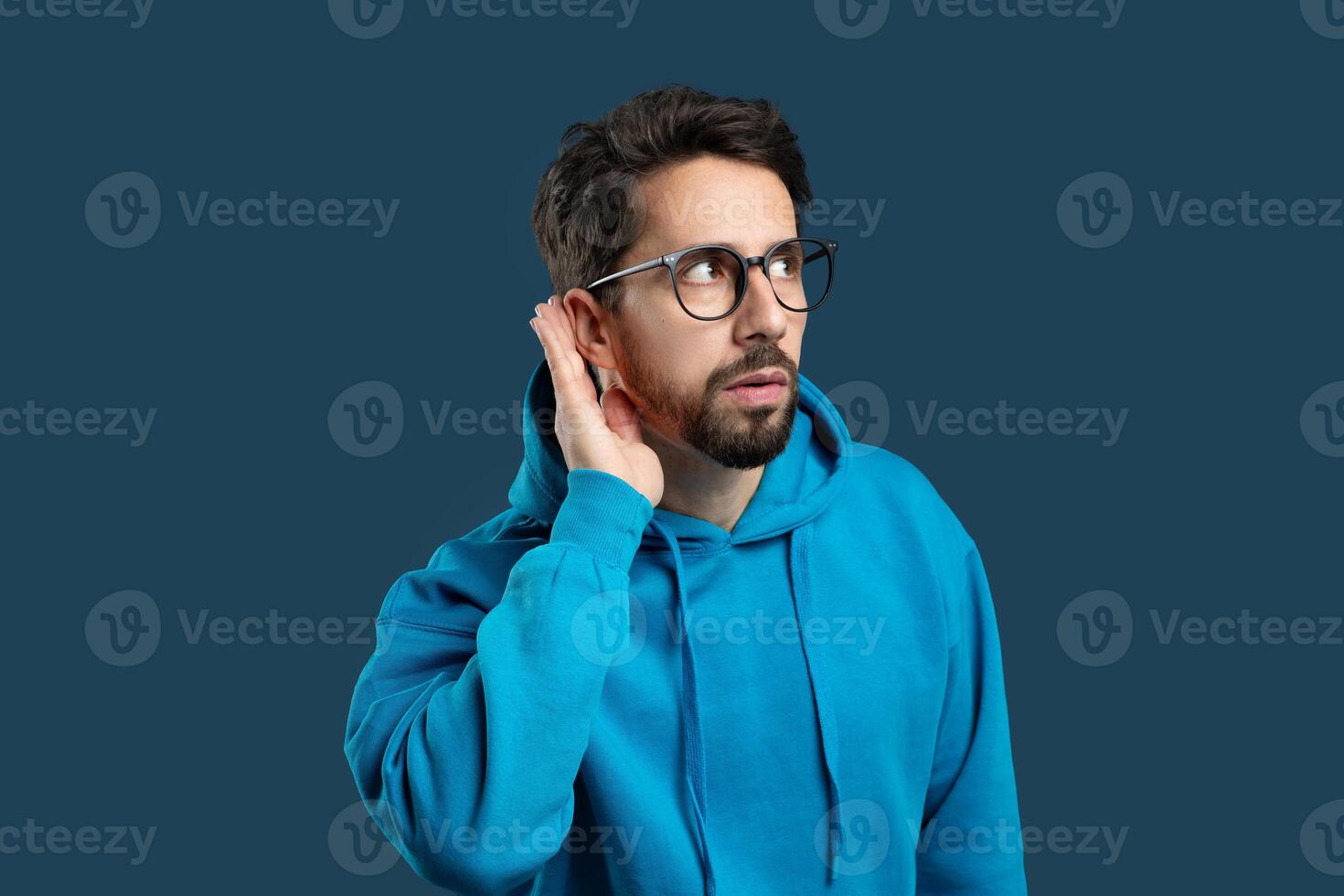 A man with a beard and glasses, dressed in a blue hoodie, stands against a solid color background. He appears focused as he cups his hand behind his ear, trying to hear something more clearly. photo