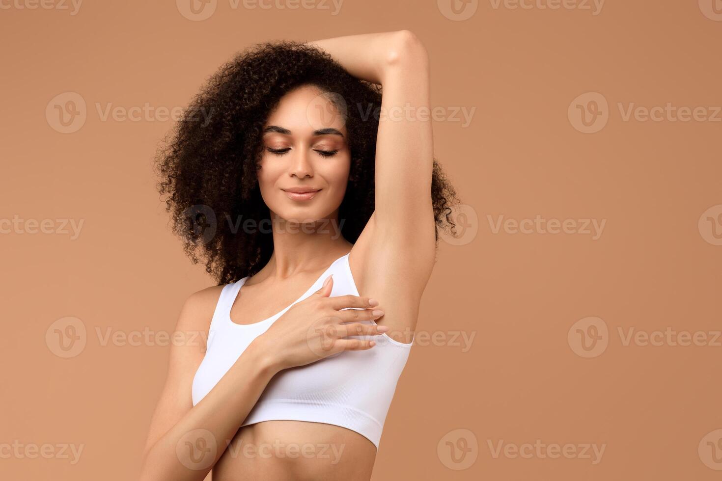 African American woman with curly hair is smiling and holding her arm up, showcasing her underarm. She is wearing a white tank top against a light brown background. photo