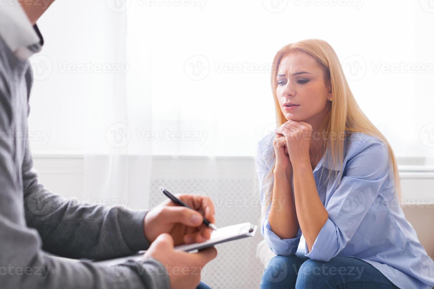 A woman sits with an expression of concern while talking to a counselor, who is taking notes on a clipboard. They are in a bright room, with the focus on the interaction and emotional support photo