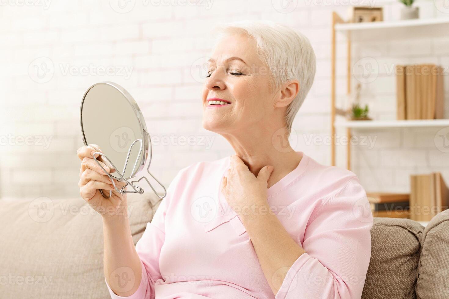 Senior woman is seated on a couch, holding a mirror in her hand. She appears to be examining her neck intently. The room is dimly lit, with soft furniture in the background. photo