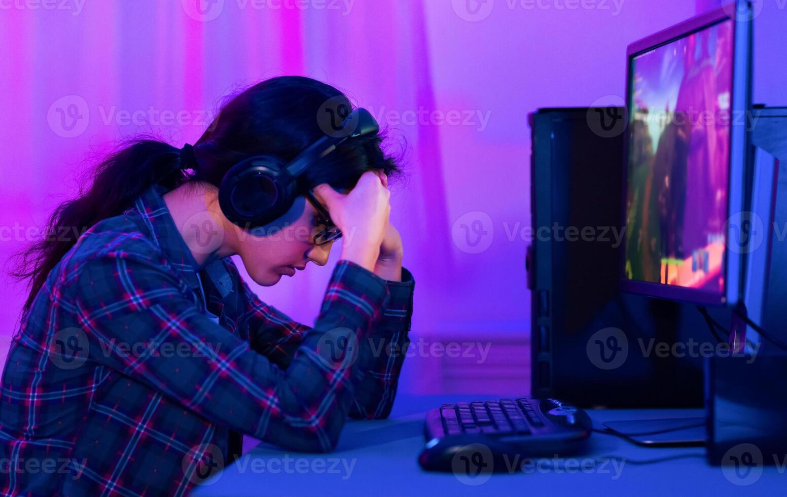 A young woman sits at a computer desk with her head in her hands, looking defeated. She is wearing headphones and glasses and is likely playing a game on her computer. photo