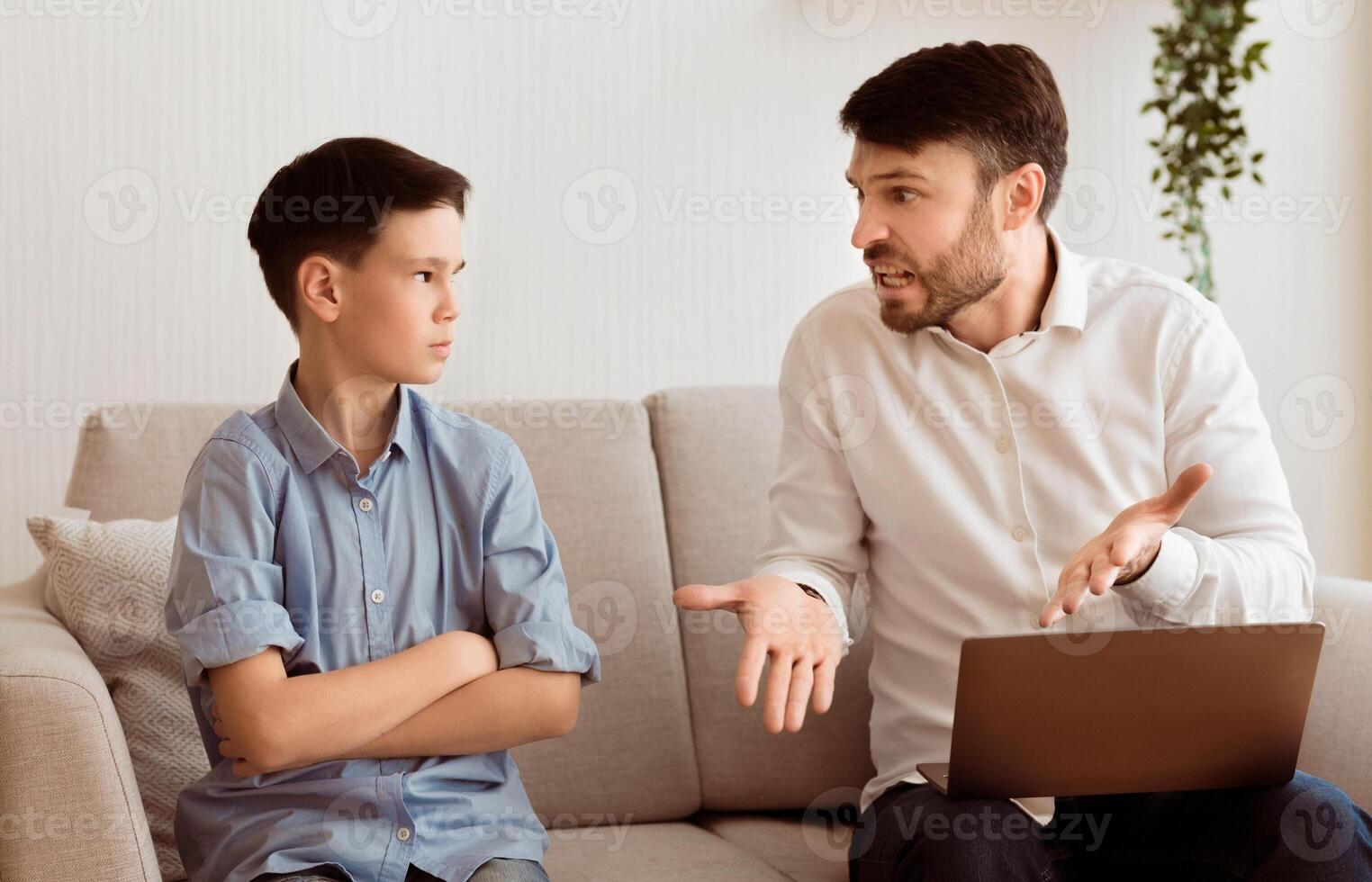 Generations Misunderstanding. Irritated Dad Yelling At Offended Son Working On Laptop Sitting On Sofa Indoor. Selective Focus photo