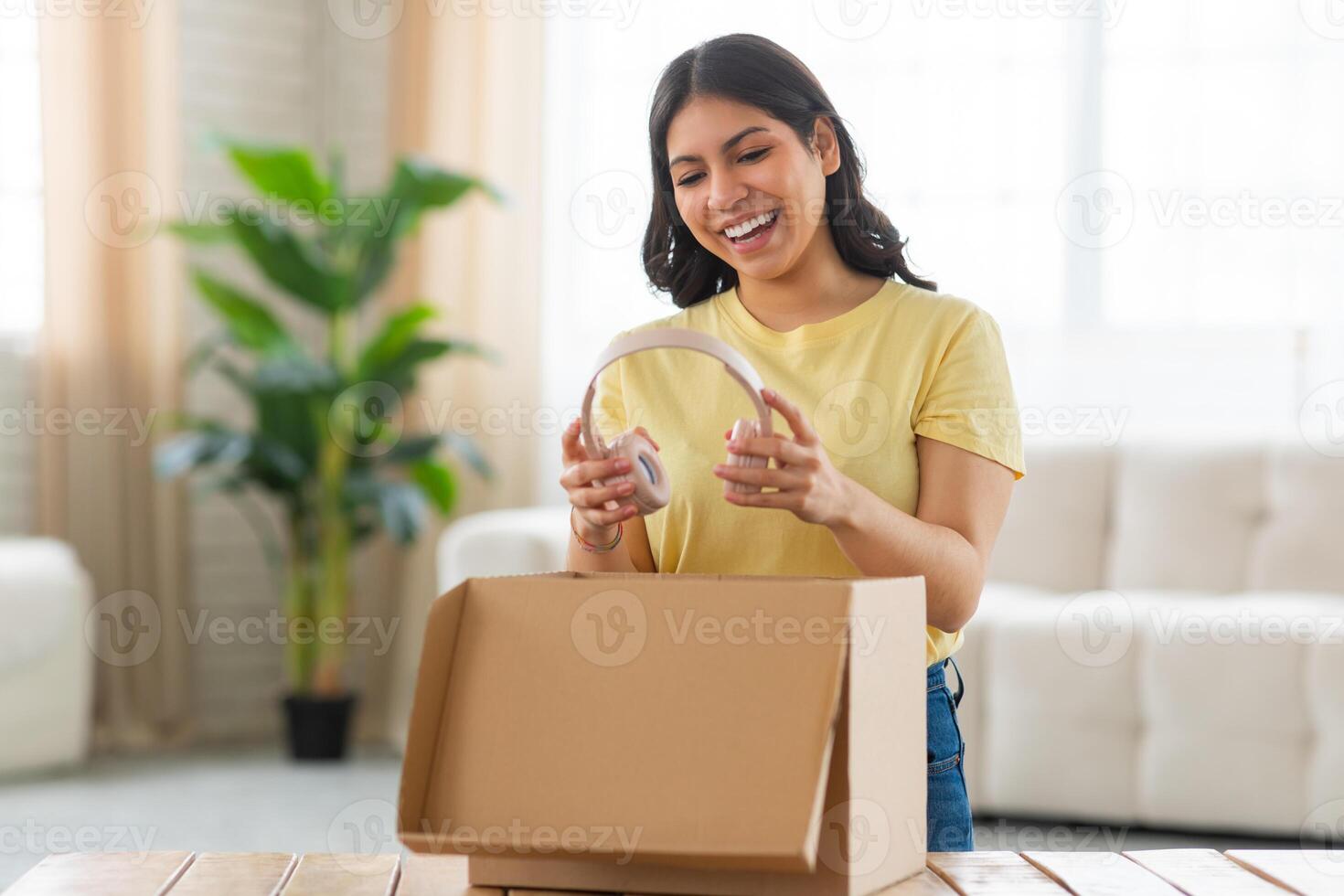 A young middle eastern woman stands in her sunlit living room, filled with excitement and a bright smile as she unpacks a cardboard box on a table, holding headphones photo
