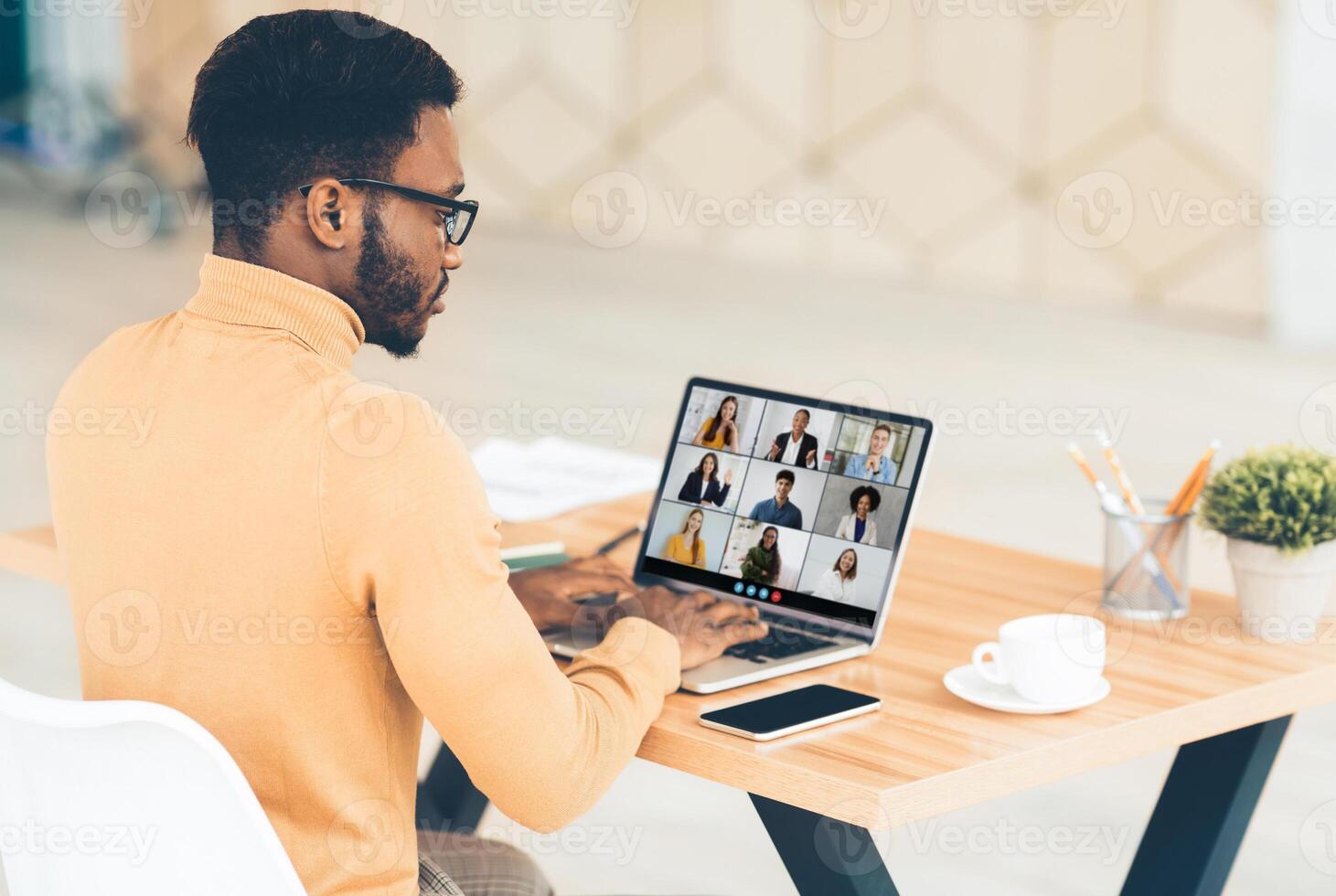 Black man wearing glasses and a light brown sweater sits at a desk, focused on his laptop screen. The screen displays a call with several participants. The workspace is clean and modern photo
