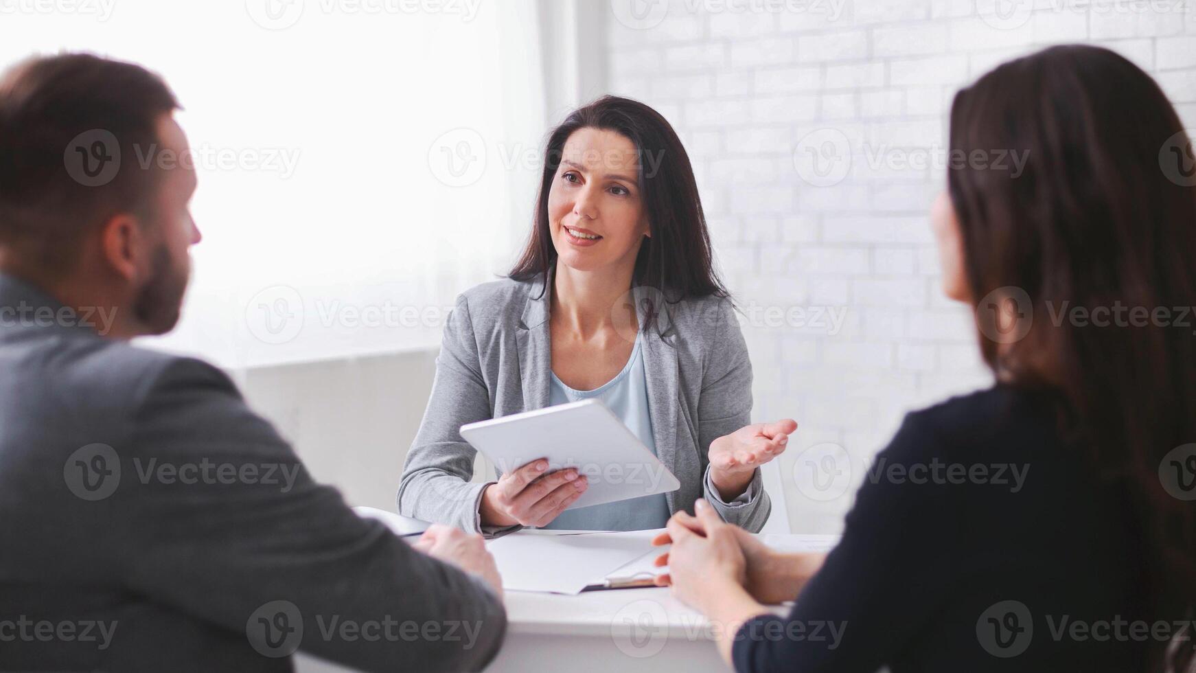 A businesswoman is presenting ideas to a couple during a business meeting. She is holding a tablet and gesturing with her hand as she speaks. The couple is listening attentively photo