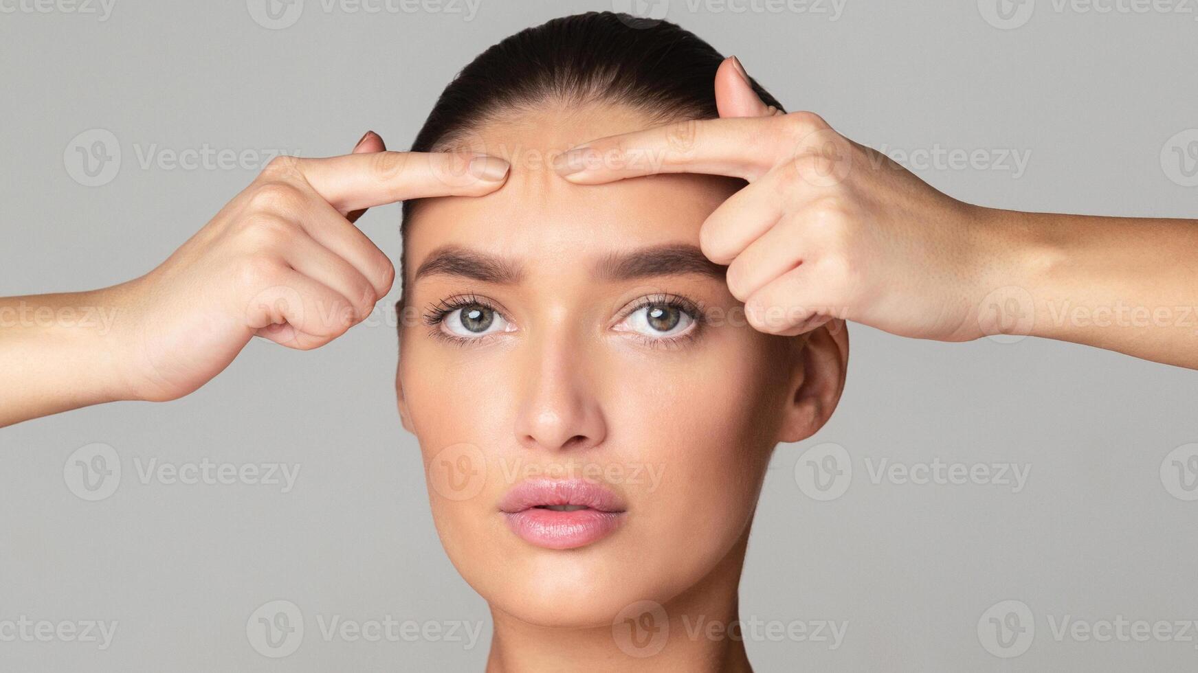 A woman with tied-back hair is using her fingers to squeeze a pimple on her forehead. The setting appears bright and indoor, suggesting a well-lit room. She has a calm yet focused expression photo