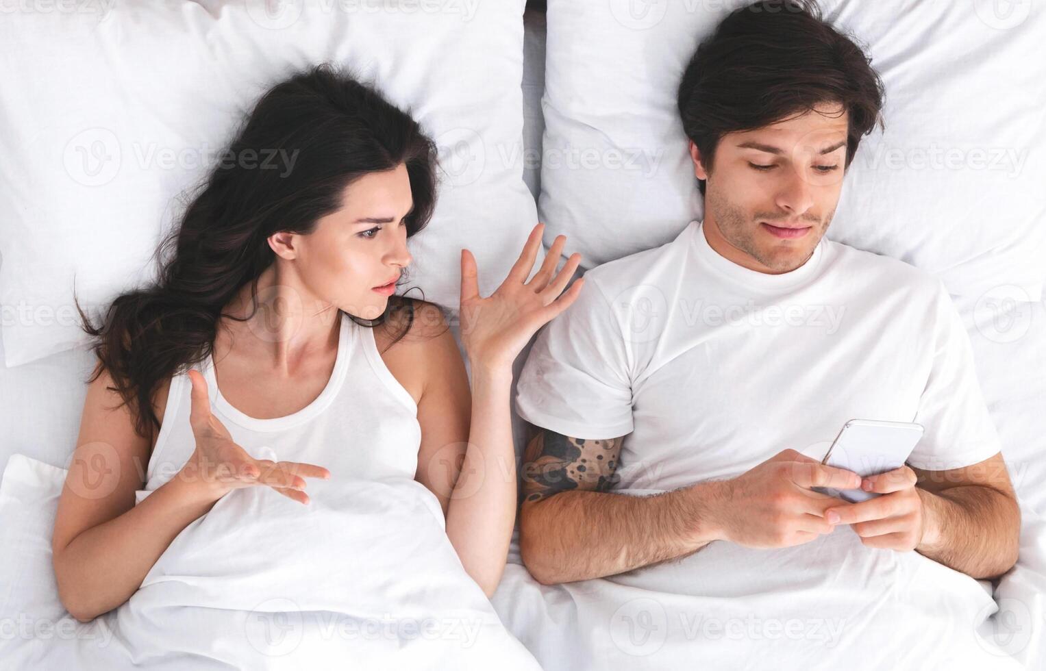 An image depicting a couple lying in bed. The woman is looking at the man, with her hand outstretched, seemingly trying to get his attention. He is ignoring her and looking at his phone photo
