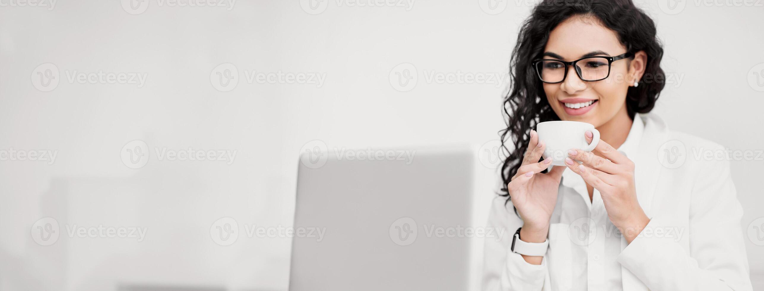 Break At Work. Happy hispanic woman drinking coffee or tea, looking at laptop, working at office. Empty space photo