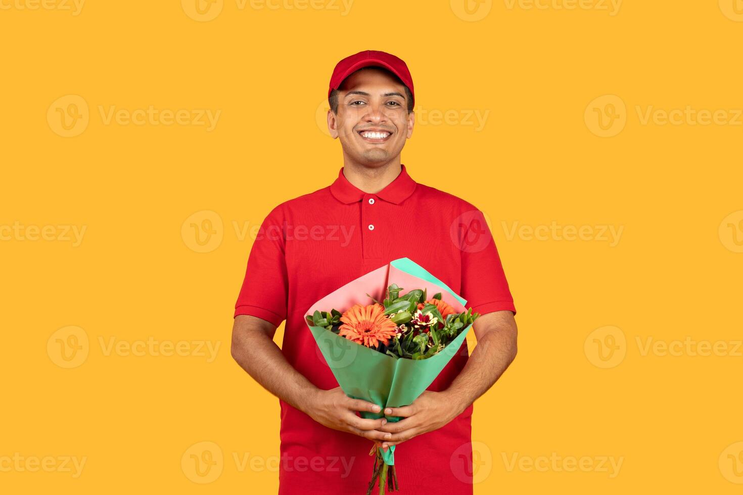 A cheerful delivery man in a red uniform holds a vibrant bouquet of flowers, radiating happiness. The yellow backdrop emphasizes the joyful atmosphere. photo