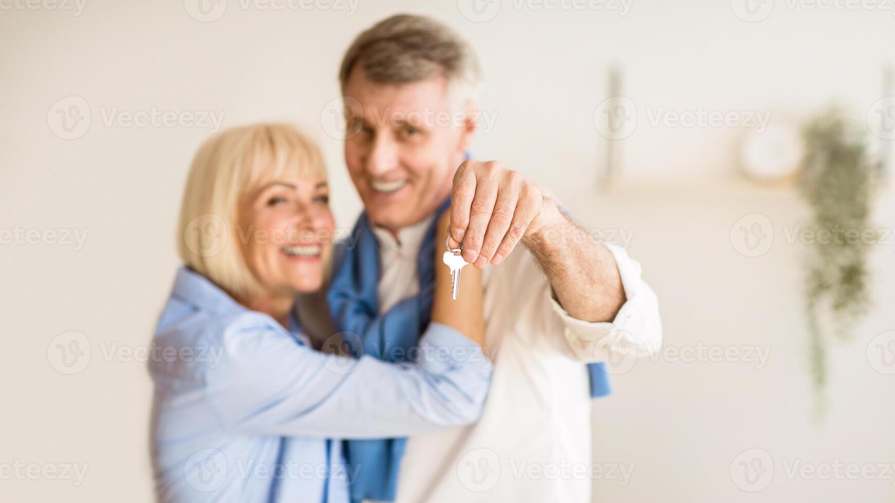 Senior couple moving into new home, man showing keys. Woman embracing husband, panorama photo