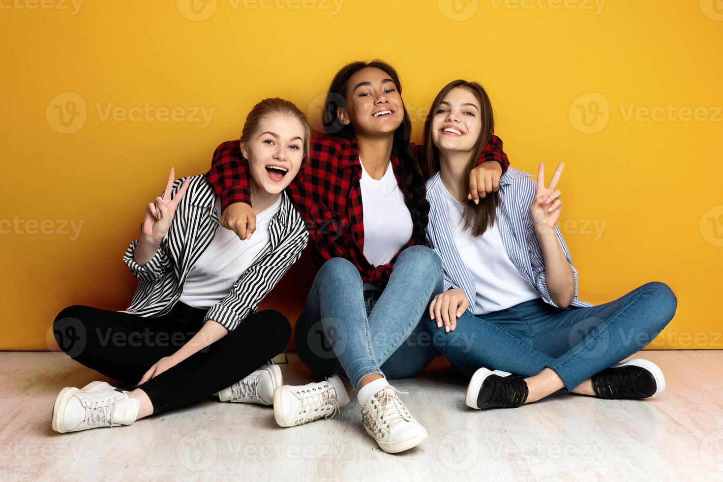Three young multiethnic girls sit closely side by side with their backs against a bright yellow wall. They are casually dressed in jeans and flannel shirts and are smiling joyfully photo