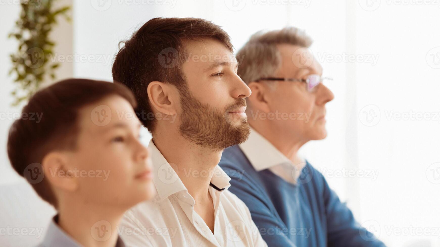 Mature man sitting between elderly father and little son looking aside indoor. Side view, selective focus, panorama photo