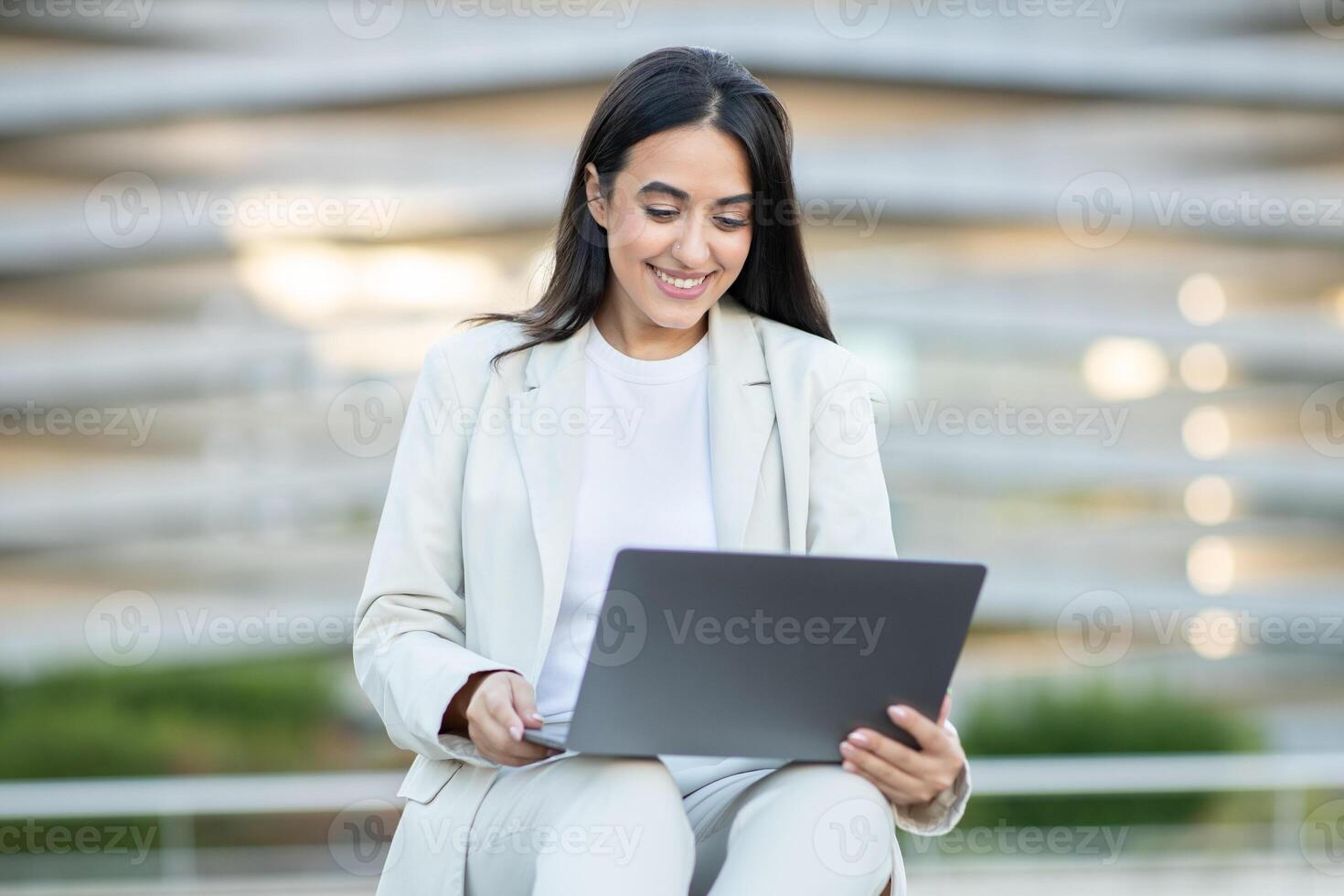 A young woman in a suit smiles while using her laptop in an outdoor environment surrounded by contemporary architecture. photo