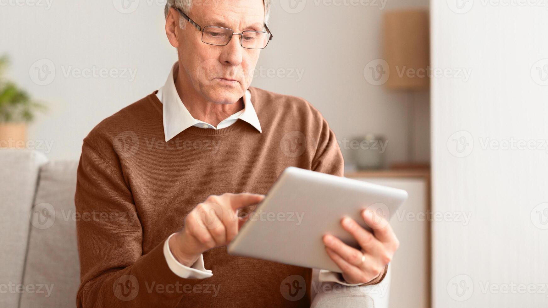 Serious Senior Gentleman Using Tablet Computer Reading News Sitting On Sofa At Home. Selective Focus, Panorama, Cropped photo