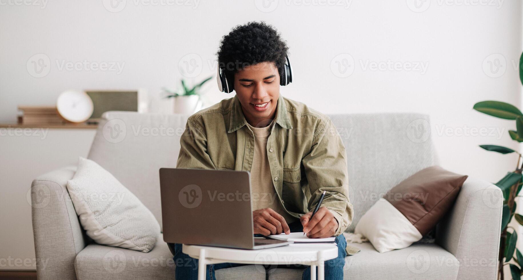 Web-based education concept. Positive teen student in headphones writing in notebook during online lecture on laptop. Cool African American guy doing homework and listening to webinar, panorama photo
