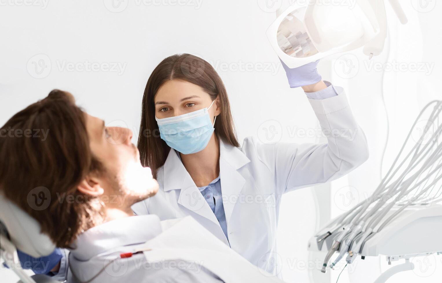 Dentist doctor woman in mask turning on lamp before making check up, patient sitting with open mouth photo