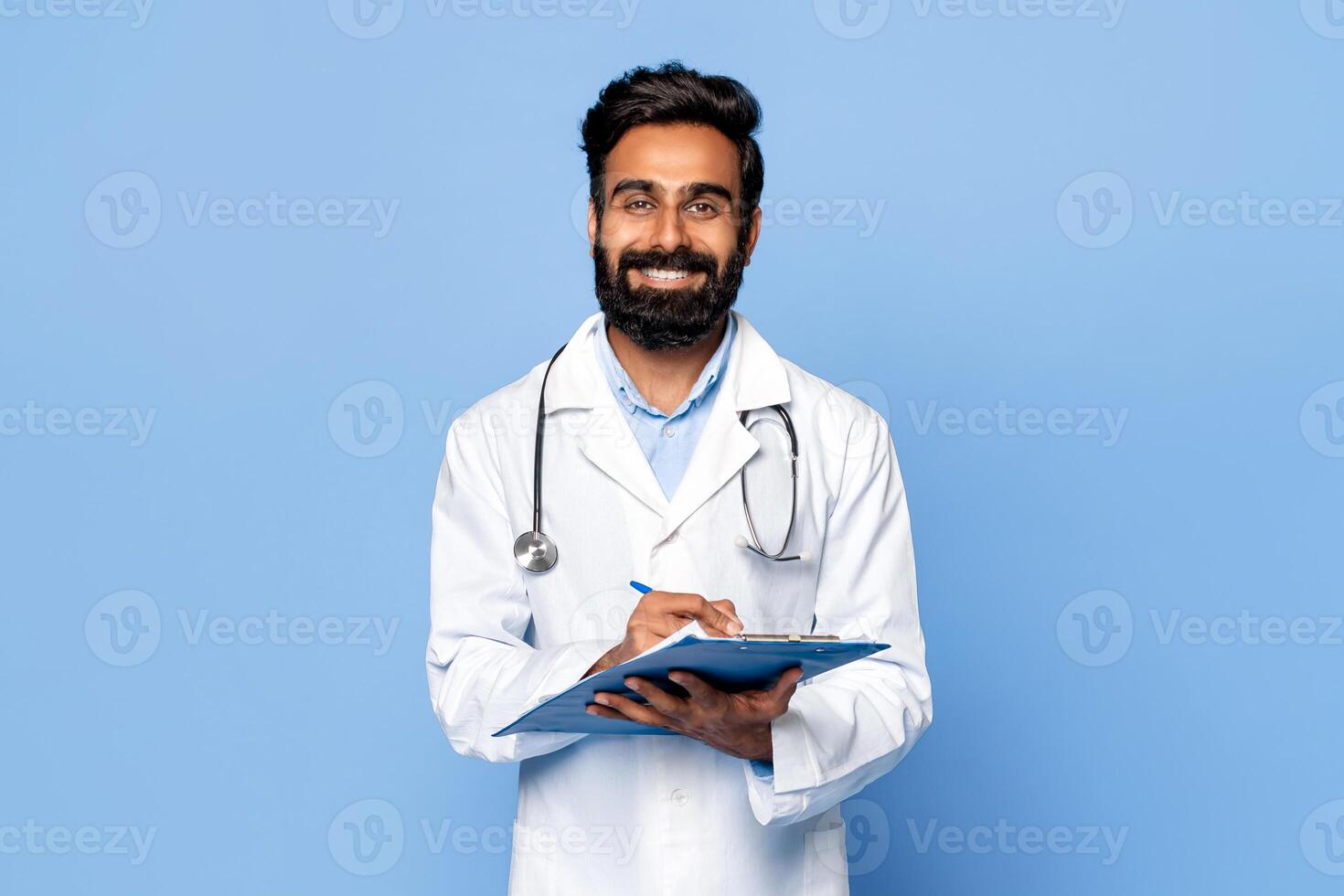 A smiling Indian man doctor with a beard is standing against a blue background. He is wearing a white lab coat and a stethoscope around his neck. He is holding a clipboard photo