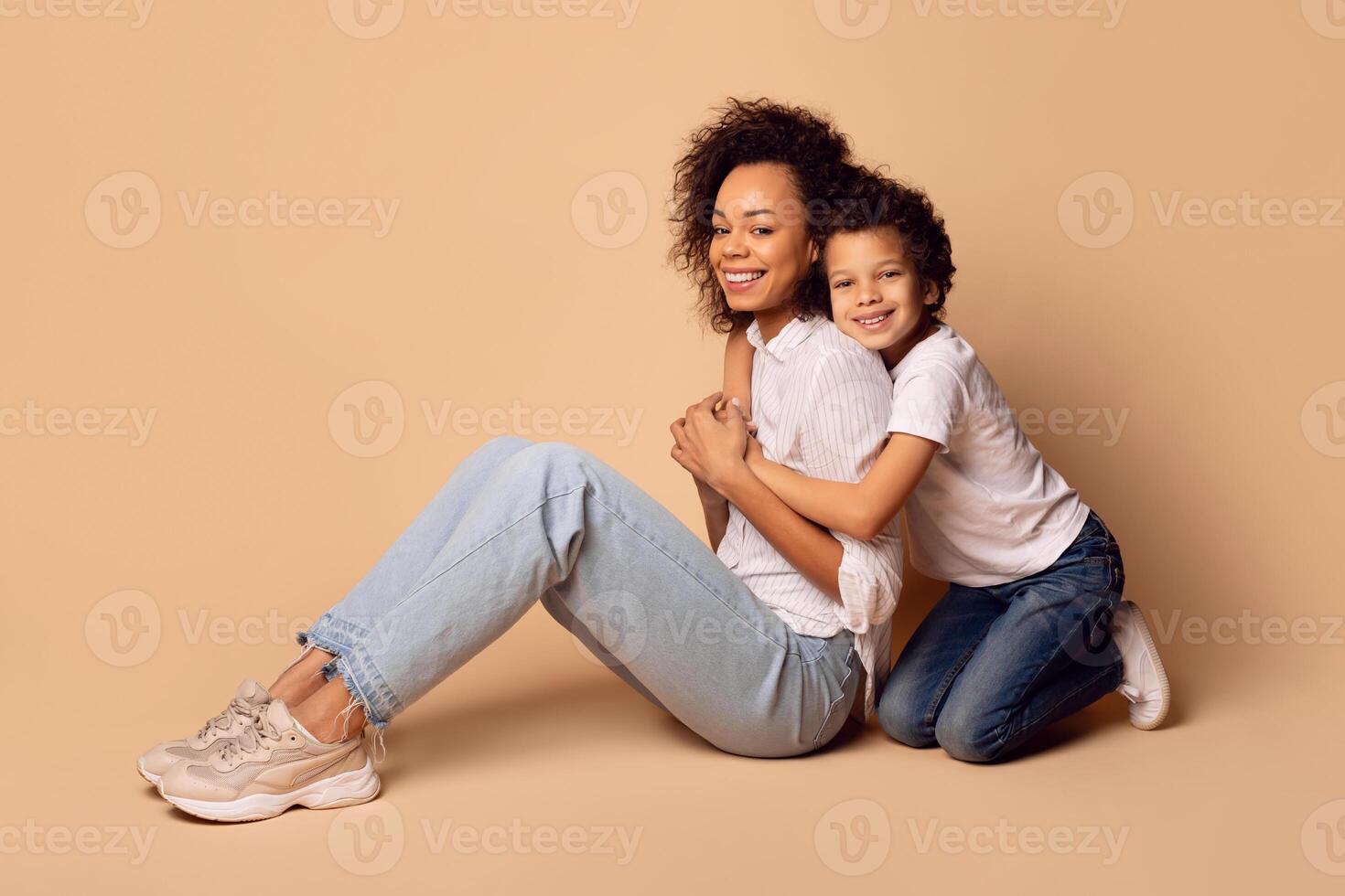 African American woman and child are seated on the ground, beside each other. They appear to be engaging in conversation or sharing a moment together. The surroundings are simple and unadorned. photo