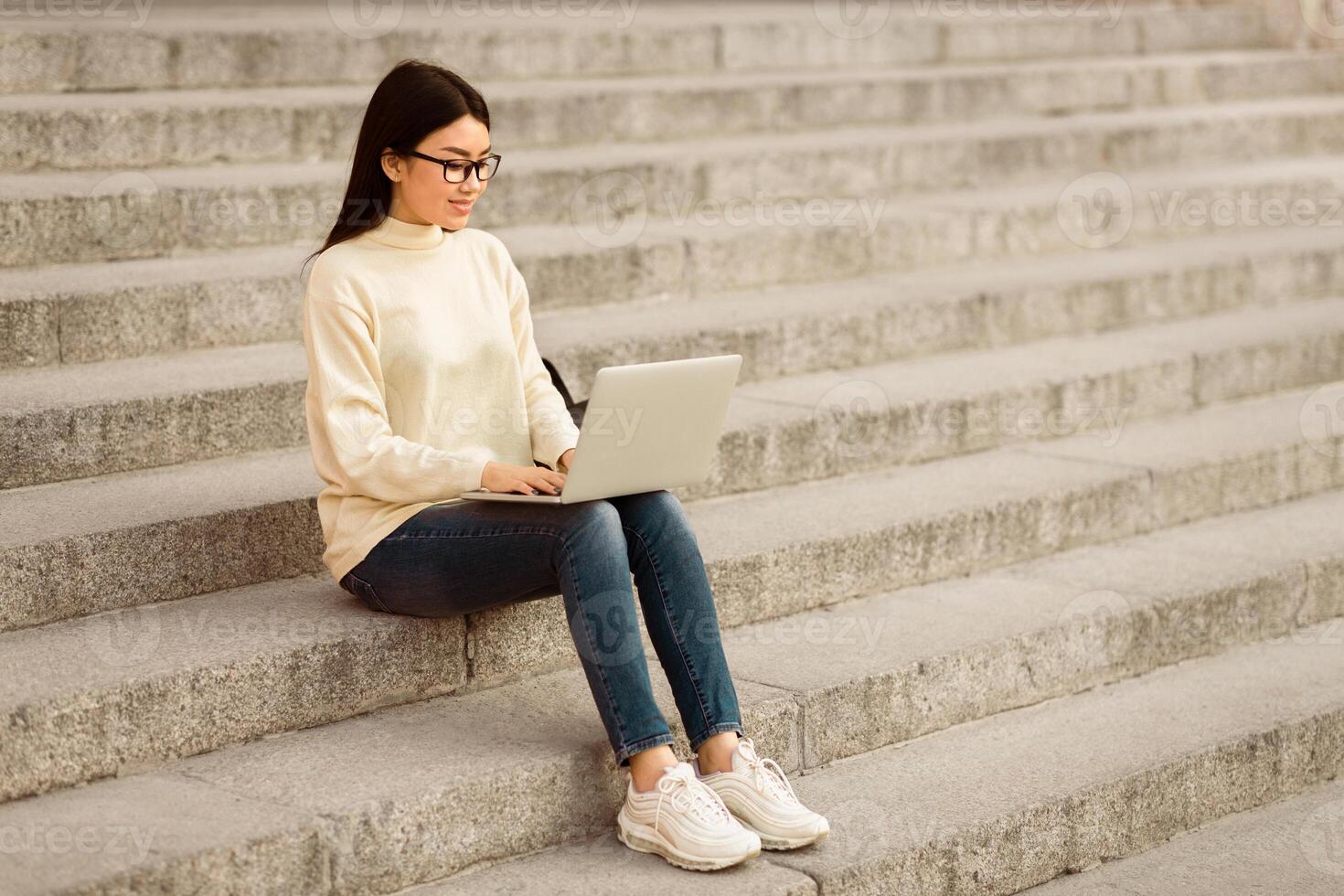 Asian woman is seated on concrete steps, typing on a laptop computer in front of her. She appears focused, with buildings and a railing in the background, copy space photo