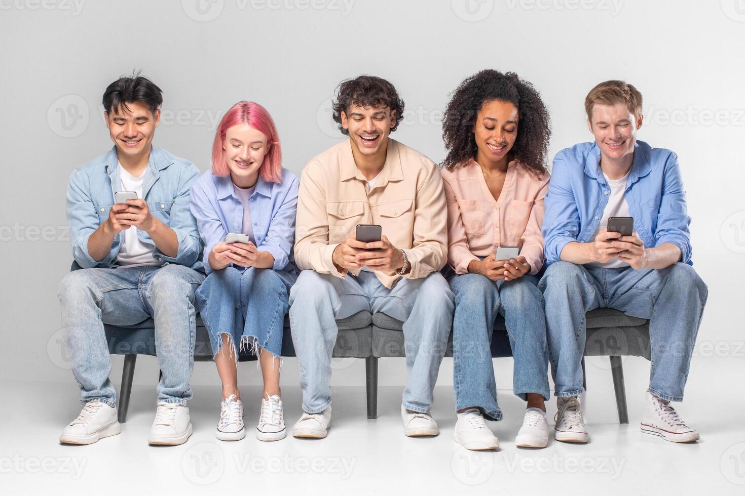 This image shows five diverse young adults sitting on a bench together. They are all looking at their smartphones. The people are casual and relaxed. photo