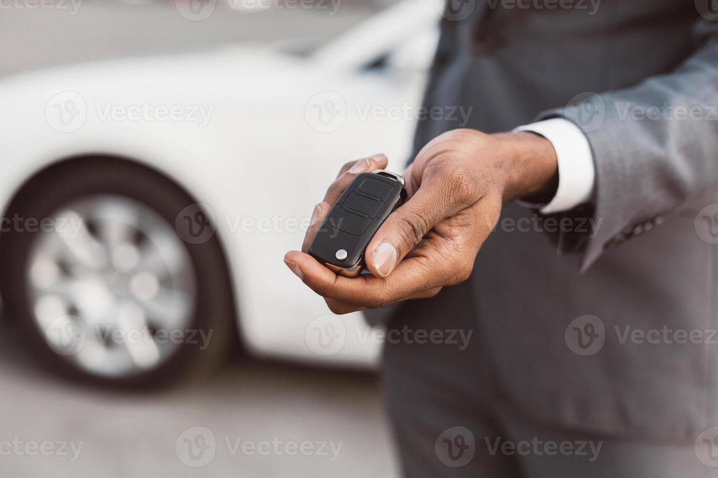 Black mans hand holding a car key in front of a white sedan. The man is wearing a gray suit and the key is a black remote. The car is partially out of focus and the mans hand is the main focus. photo